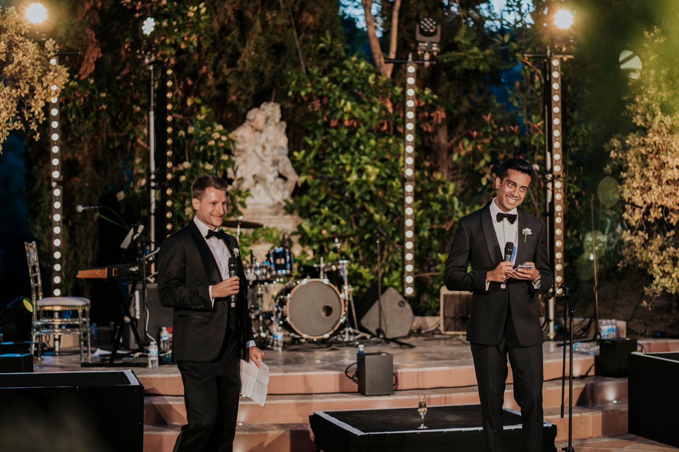 Two performers in black tuxedos sing on stage at an elegant outdoor event with sparkling lights and greenery.