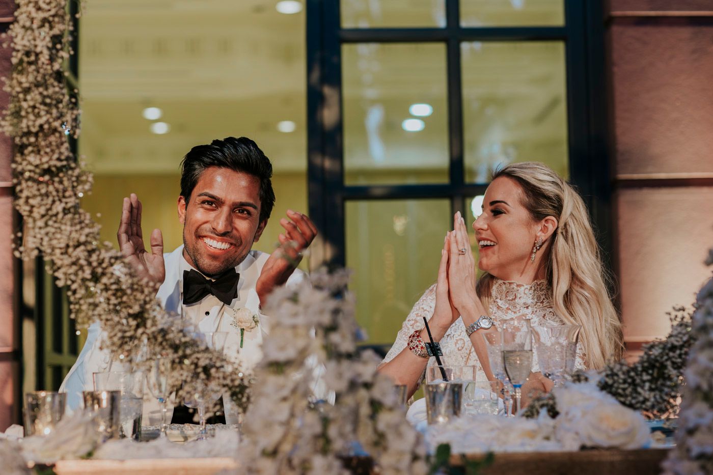 Newlyweds share a joyful laugh together at their wedding reception table adorned with floral centerpieces.