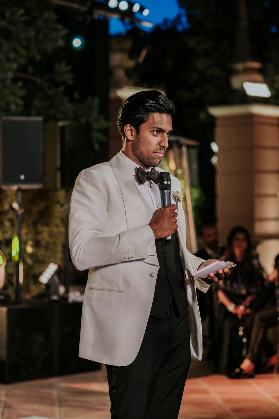 Evening wedding speech being delivered by someone in a white dinner jacket holding a microphone at an outdoor reception.
