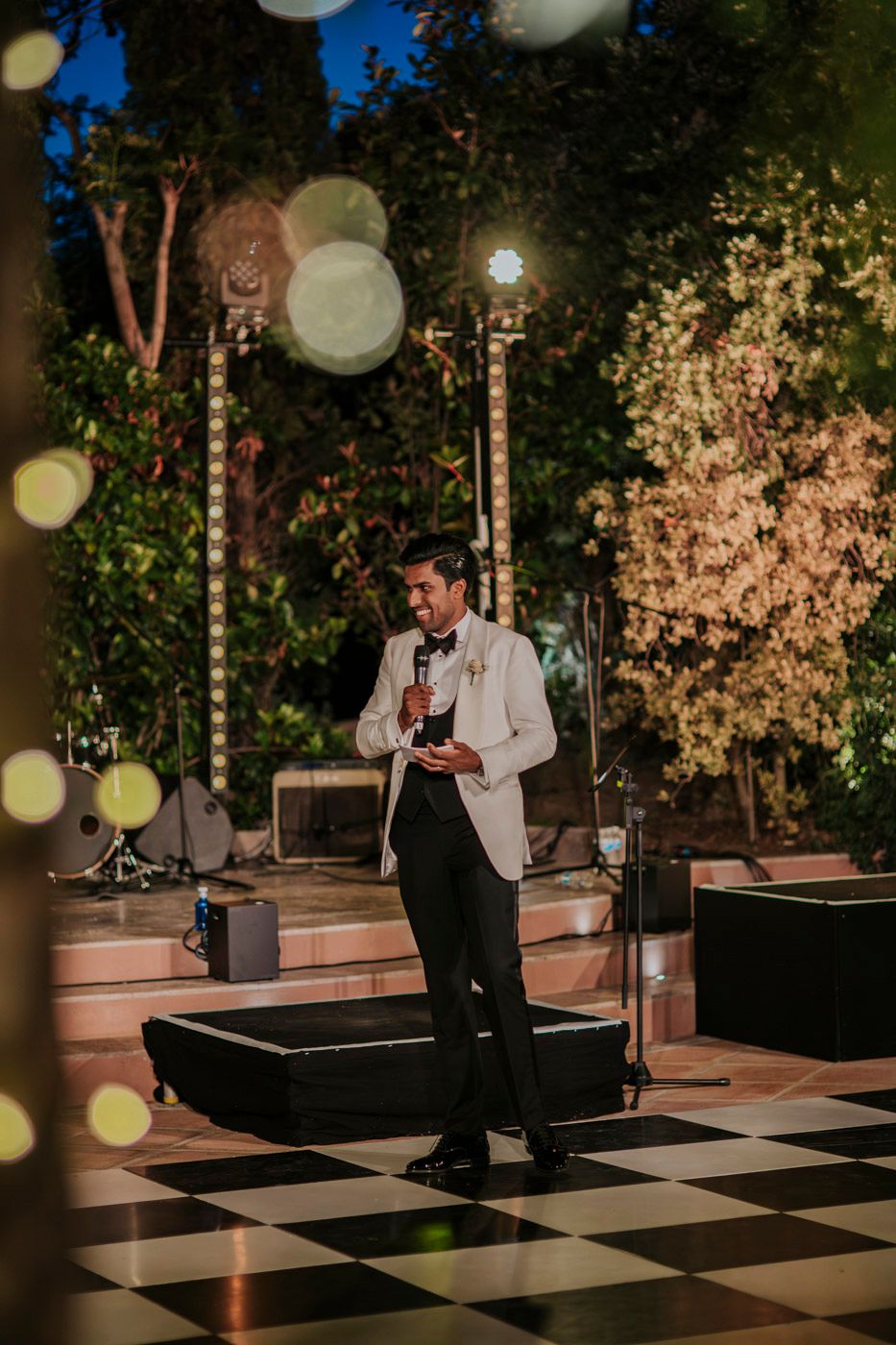 Someone in a white dinner jacket gives a speech on a checkered dance floor at an elegant outdoor evening event under string lights.