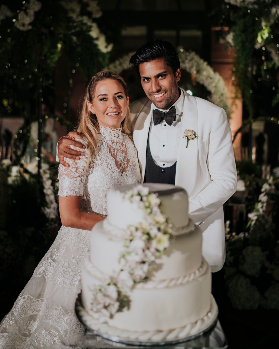 A couple stands behind their elegant white wedding cake decorated with floral accents during an evening reception.