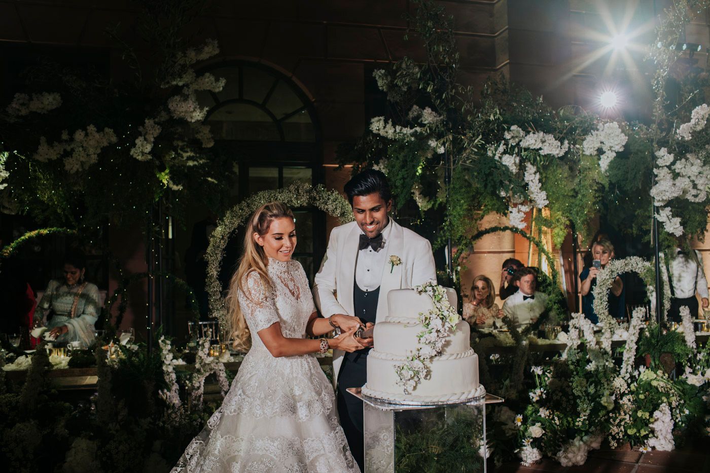 Romantic outdoor wedding ceremony at night surrounded by white floral arrangements and twinkling lights in garden setting.