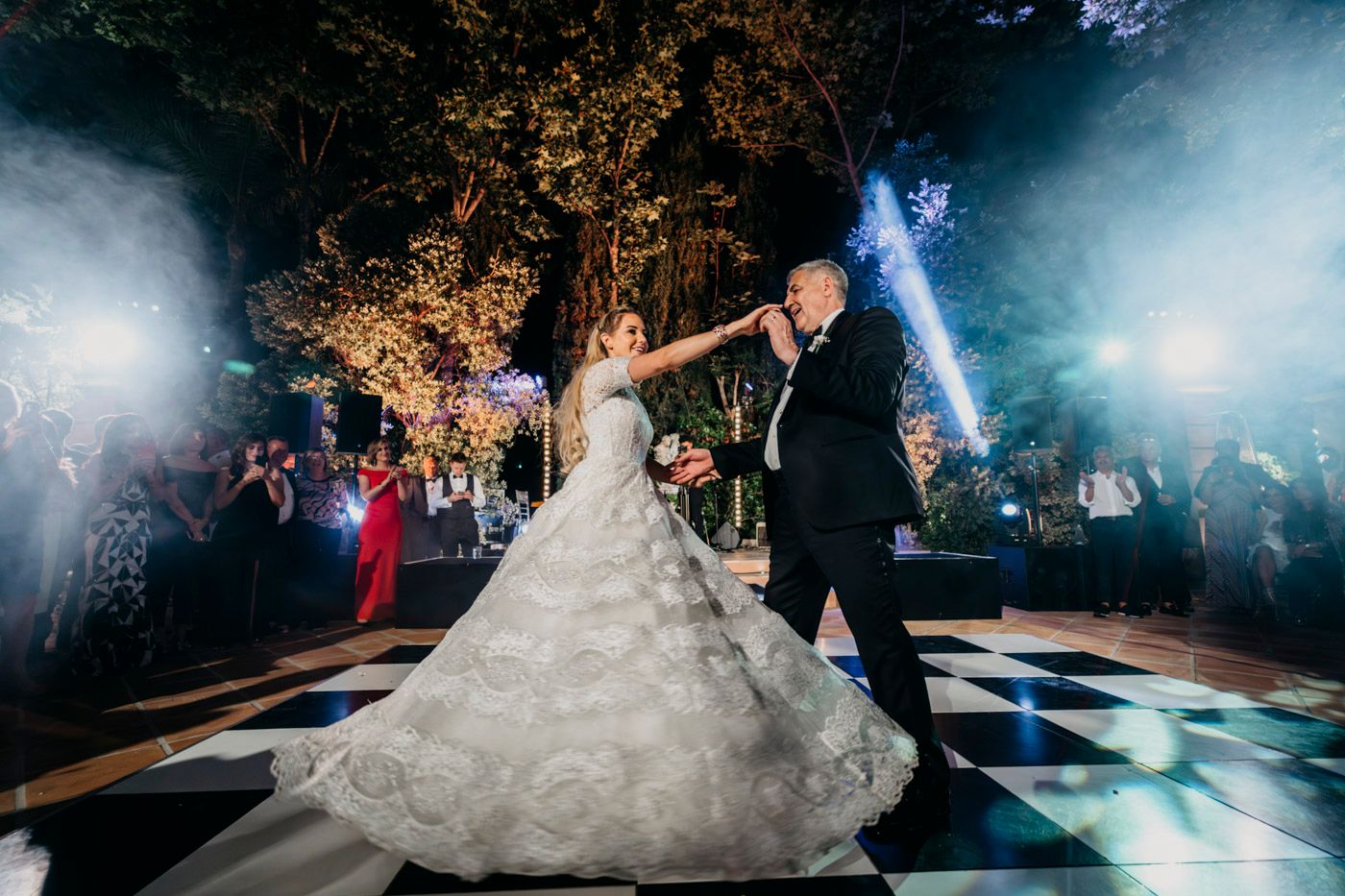 A couple's first dance on a checkered dance floor at an outdoor wedding reception with dramatic blue lighting and fog effects.