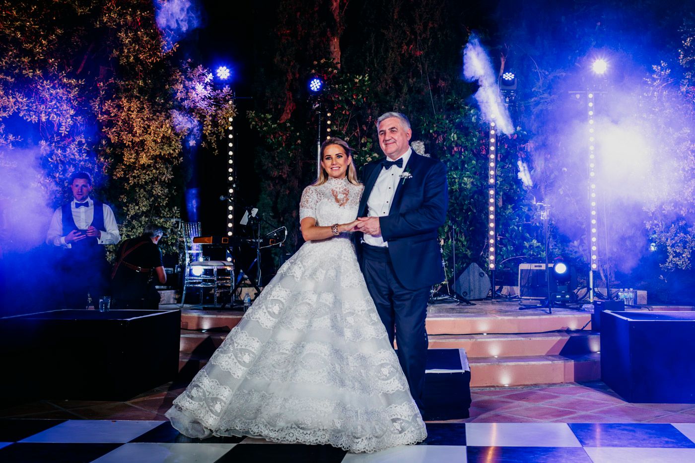 A couple dressed in formal wedding attire stand on an illuminated dance floor with blue lights and decor in the background.