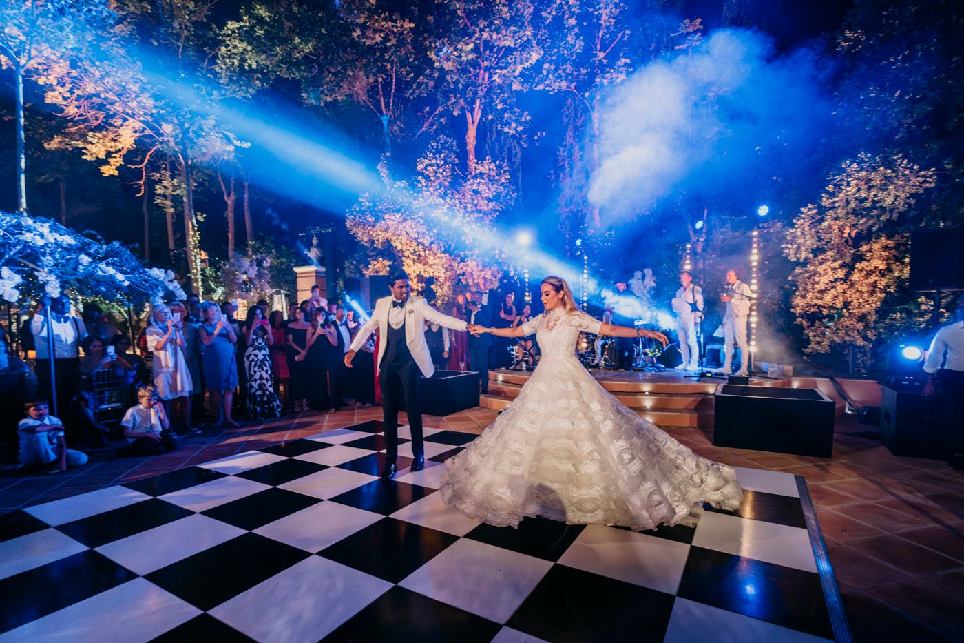 Dramatic nighttime dance on checkered floor with blue lighting and illuminated trees in background.