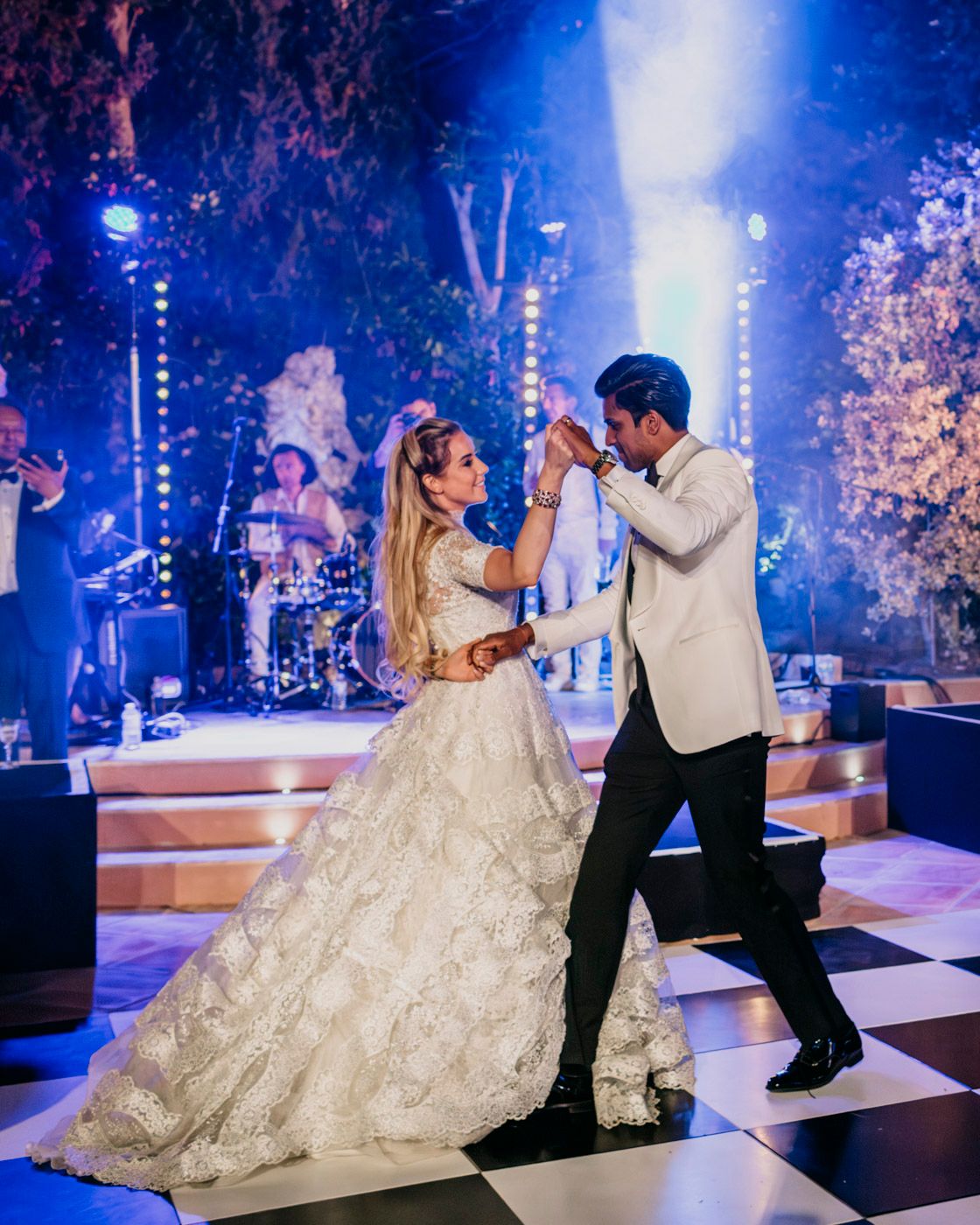 Dancing on a black and white checkerboard floor under blue stage lights and cherry blossom trees at an elegant reception.