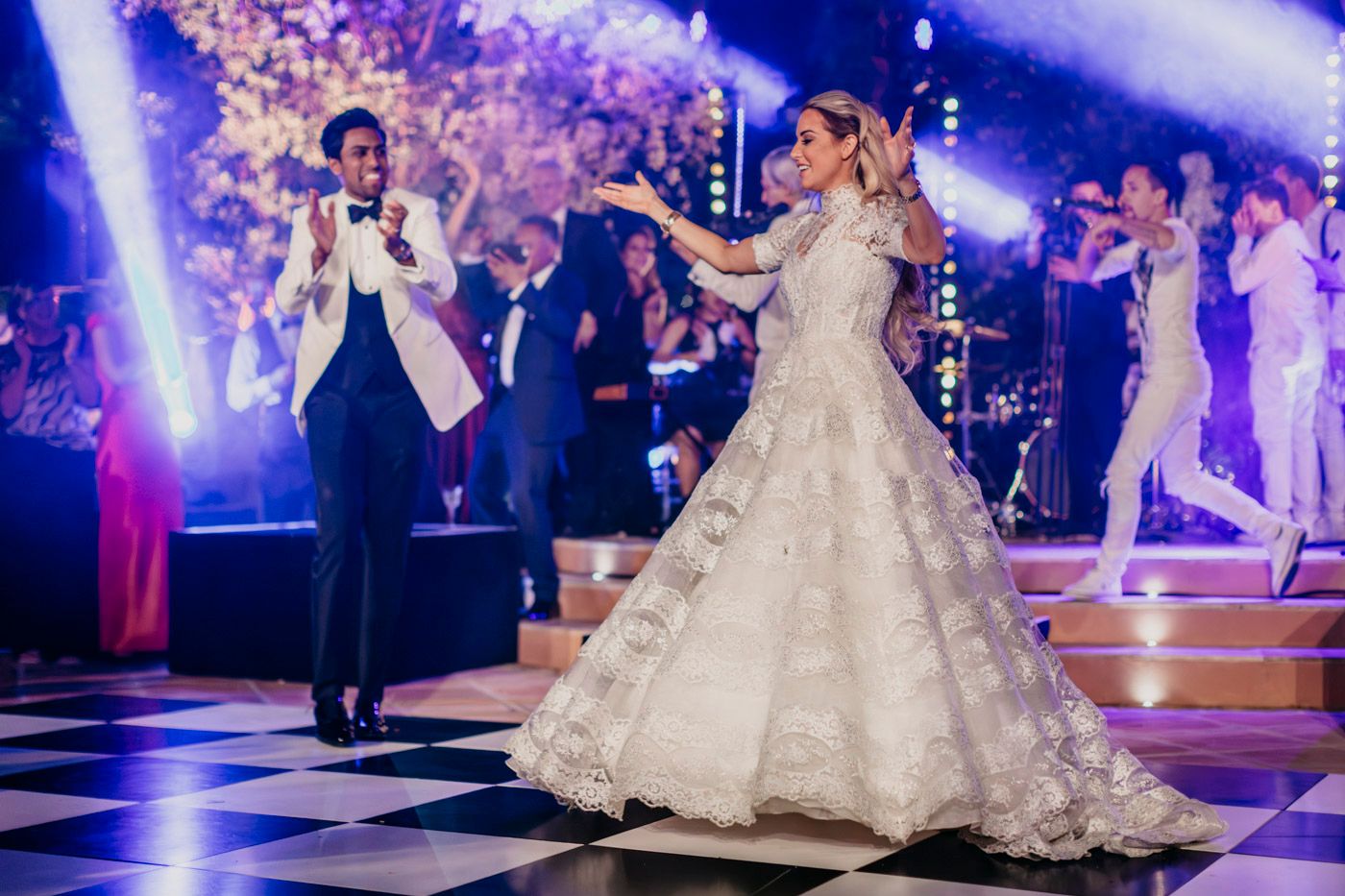 Two people dance on an elegant illuminated reception floor with blue lighting and guests visible in the background.