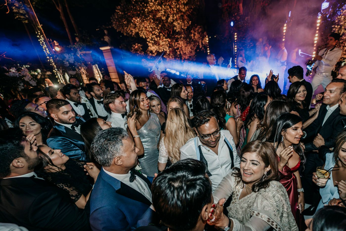 A packed dance floor full of wedding guests dancing and celebrating at an evening reception with blue lighting.