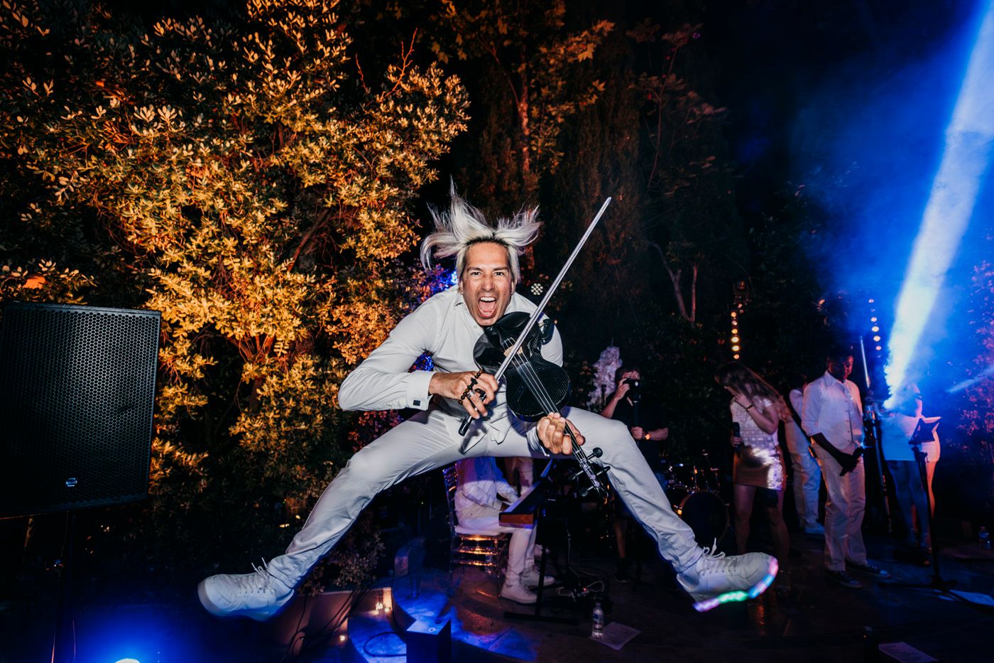 Performer in white jumpsuit and crown jumps energetically on stage with orange autumn trees and blue lighting in background.