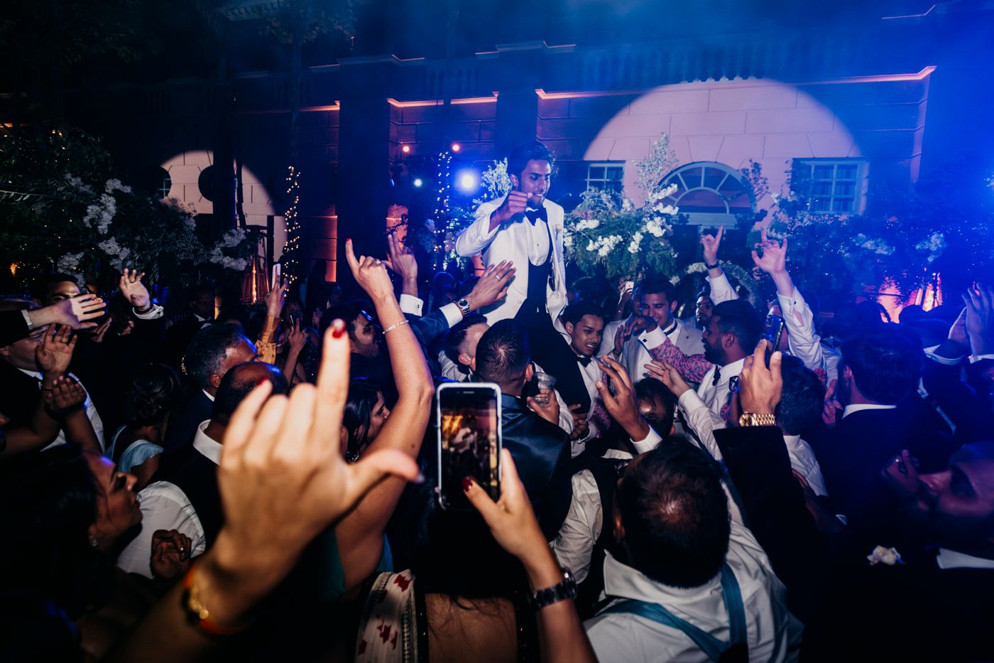 Concert crowd holds up phones to record performance on stage with blue lighting and palm tree decorations in nightclub setting.