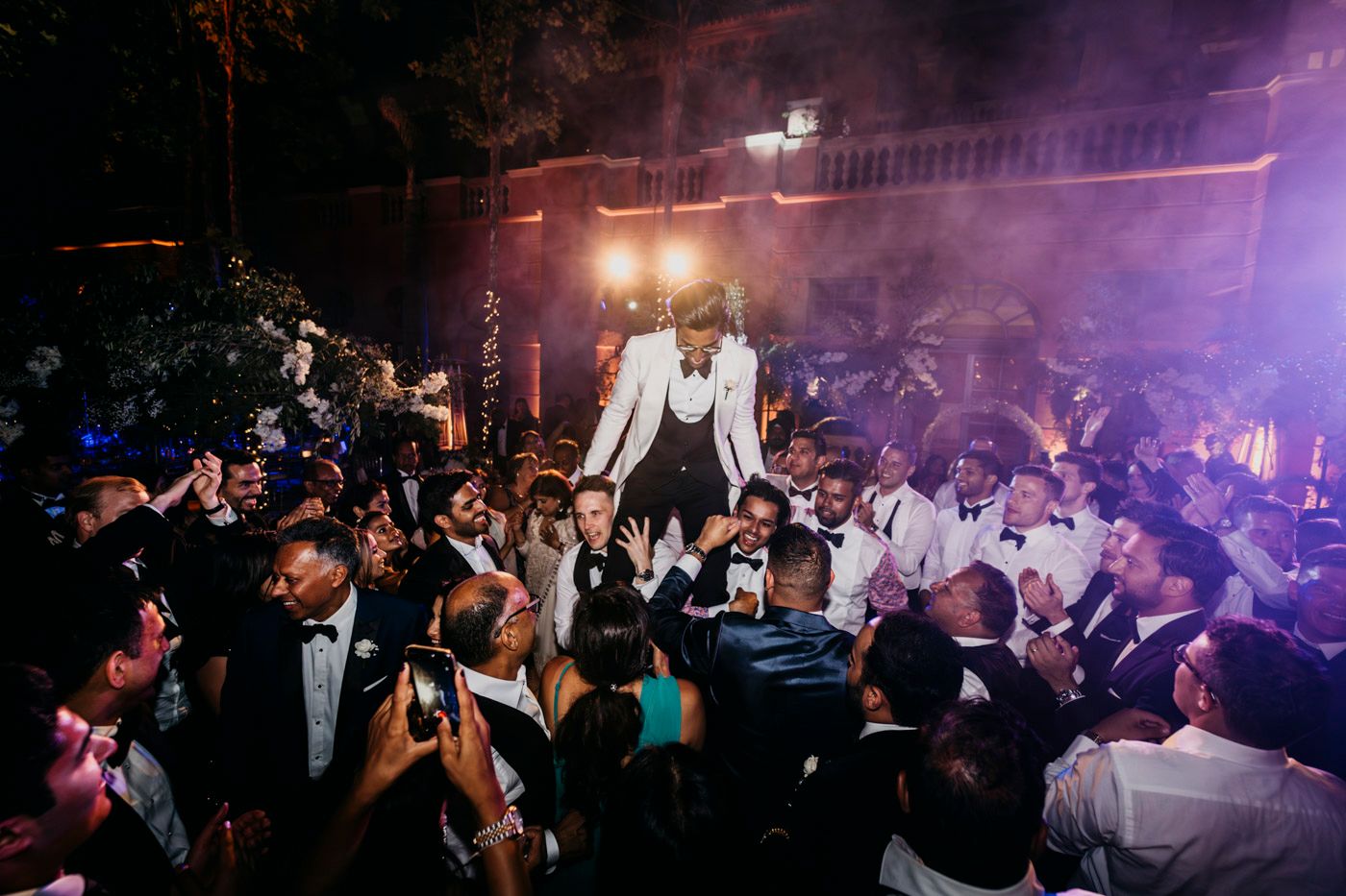 Energetic crowd at a nightclub party with someone standing elevated above the dance floor in a white suit jacket.