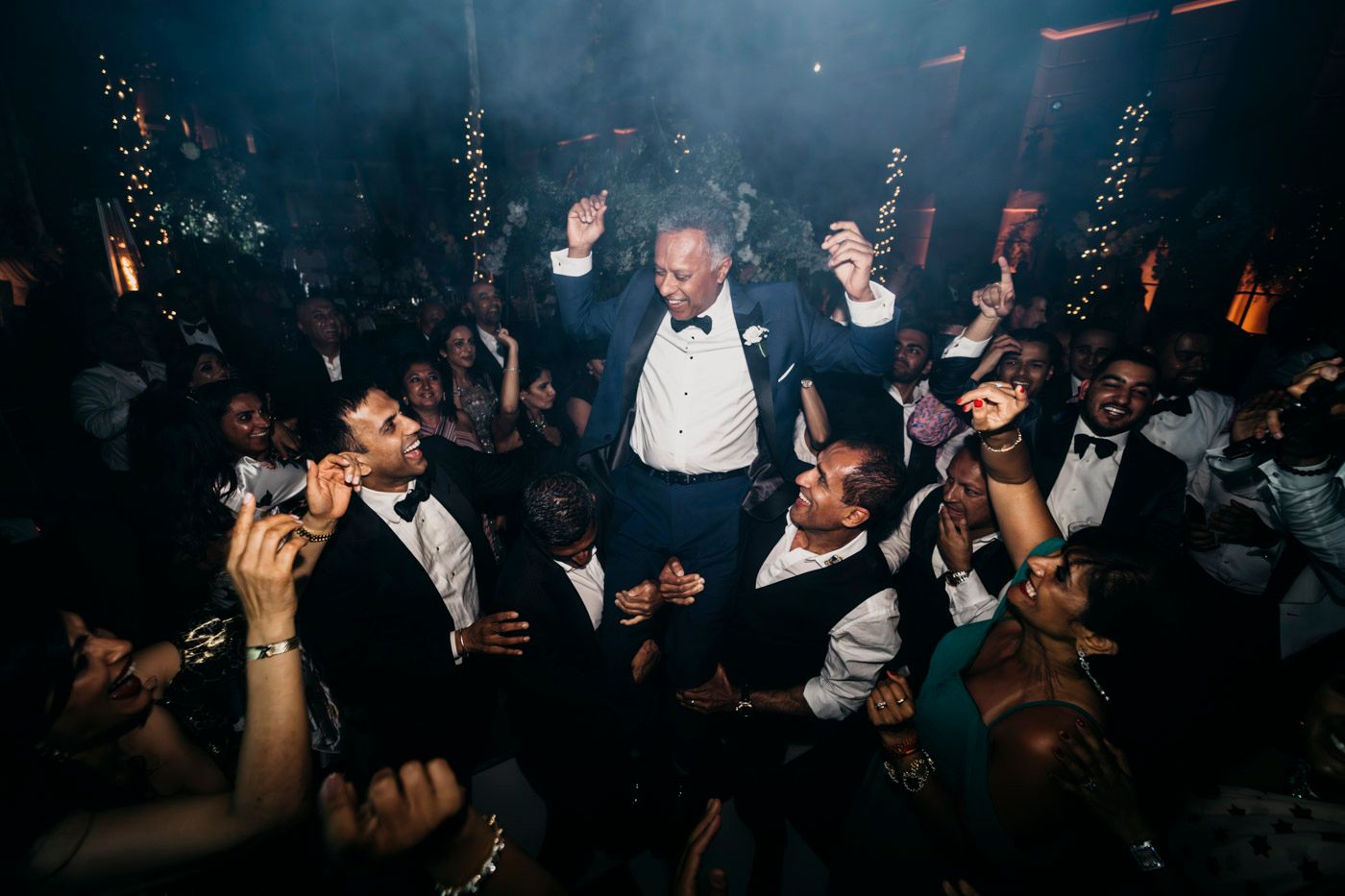 A joyous crowd celebrates at a formal nightclub party with guests dancing and raising their hands in the dark atmosphere.