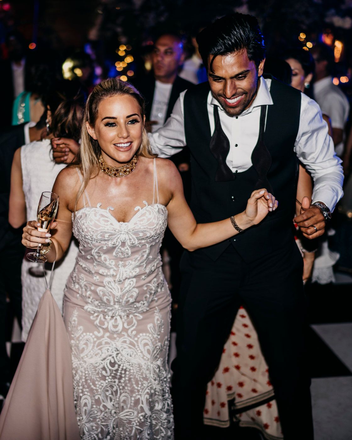 A joyful bride in a lace wedding dress dances with a guest in a vest at an evening reception party.