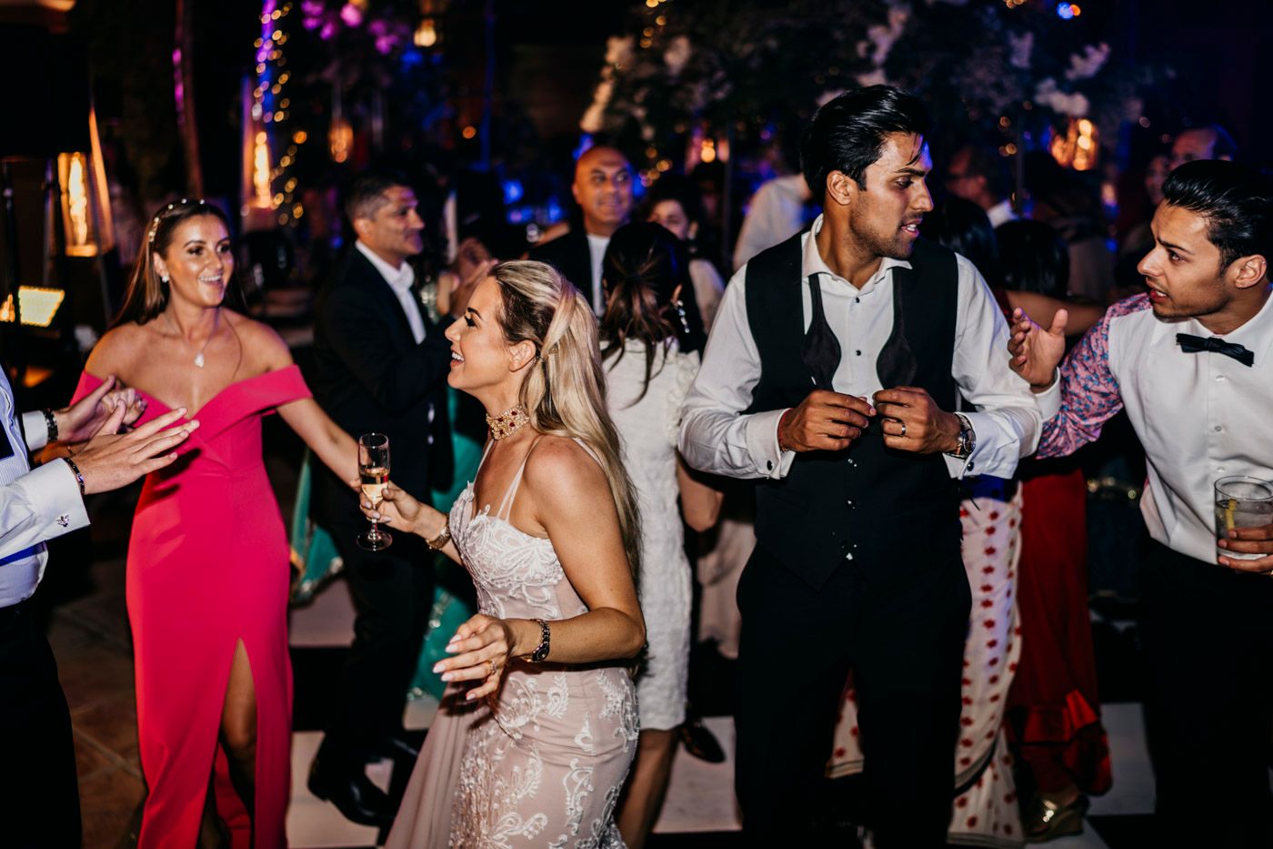 Wedding guests dancing together at a reception with colorful lights in the background, creating a vibrant party atmosphere.