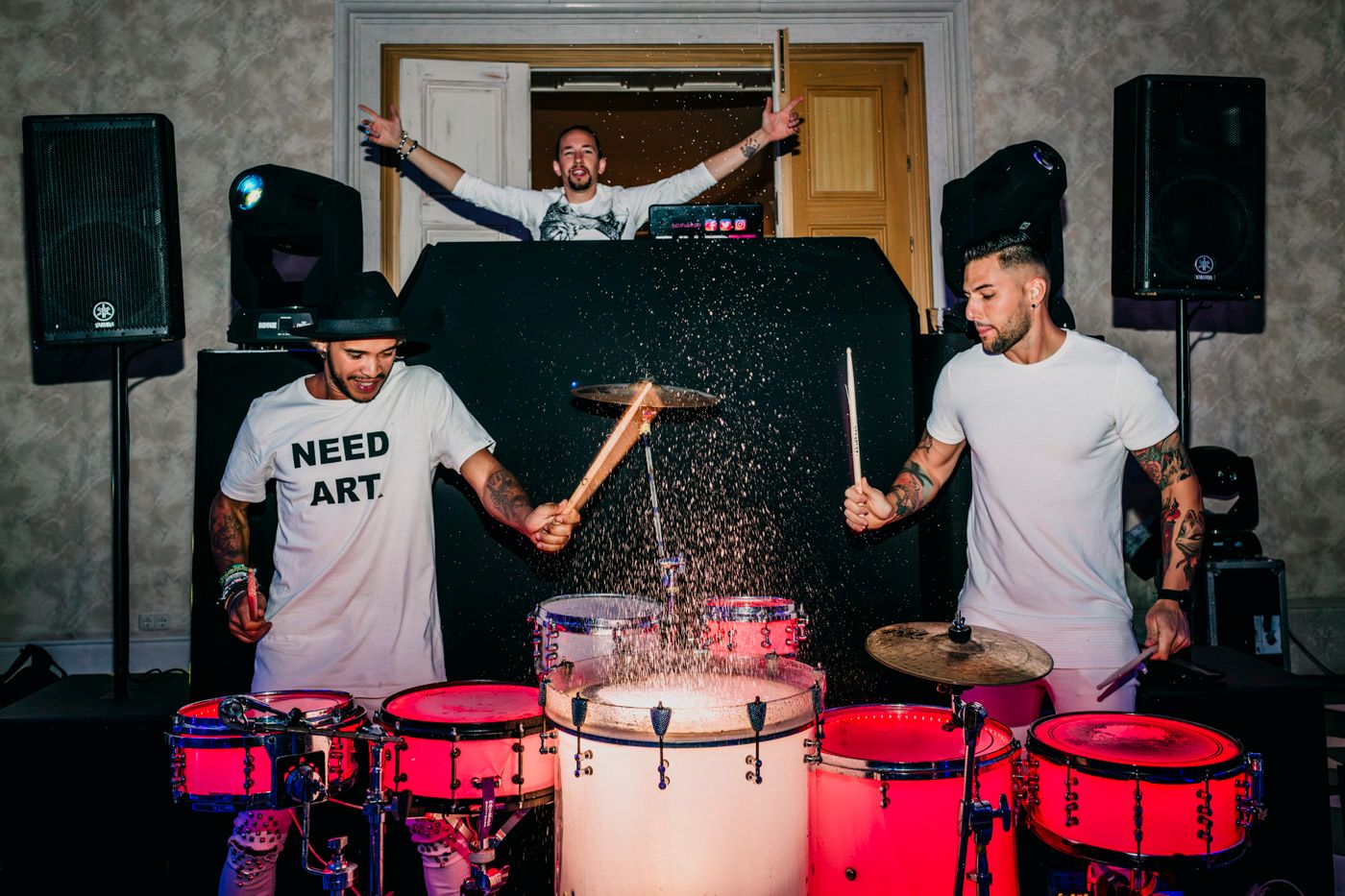 Drummers in white shirts perform with pink LED drums while DJ plays on elevated platform behind dark table.