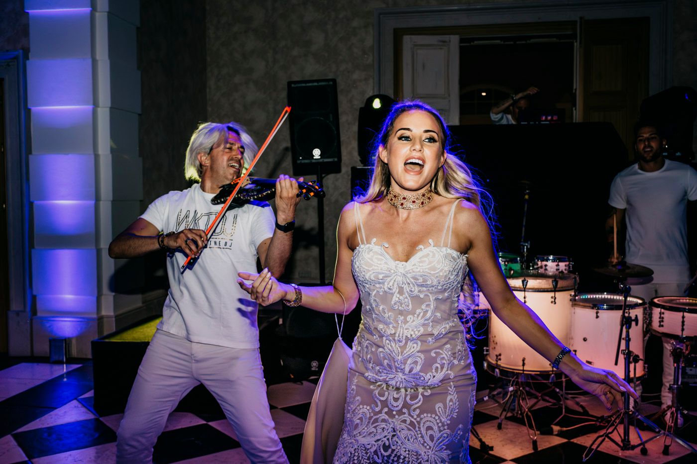 A bride in a sparkly dress dances energetically at a reception while someone poses with drumsticks behind her.