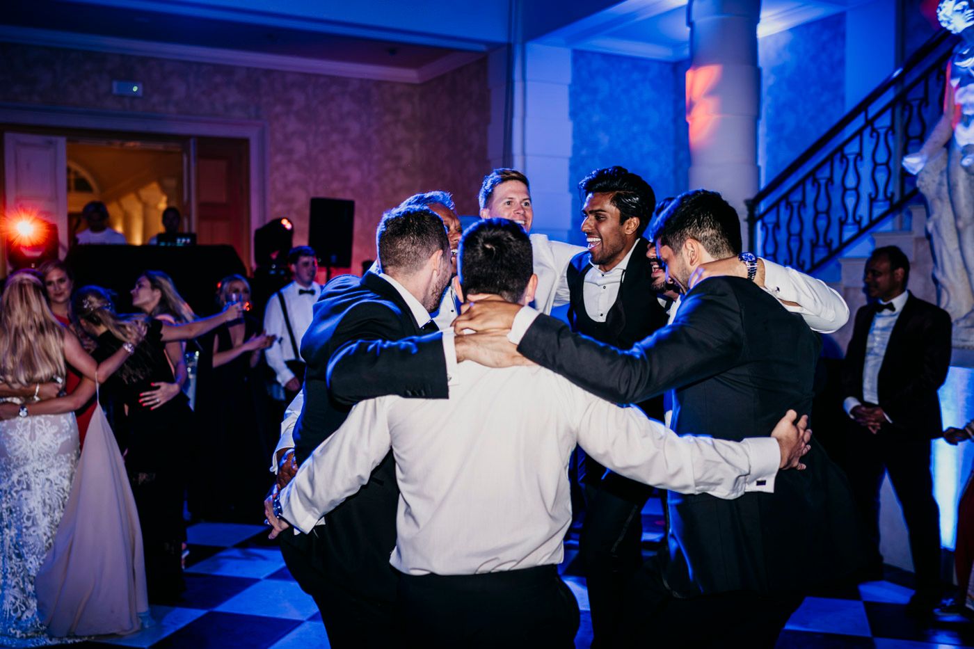 Groomsmen celebrate on the dance floor at a wedding reception with blue lighting in the background.