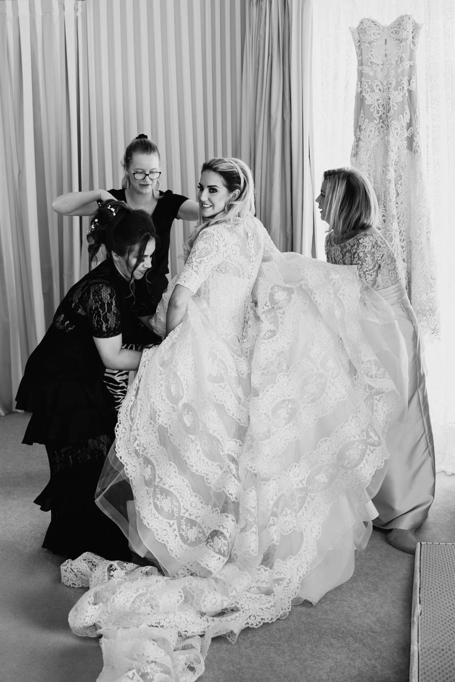 Behind-the-scenes black and white photo of a bride getting dressed in an ornate lace wedding gown with assistants helping.