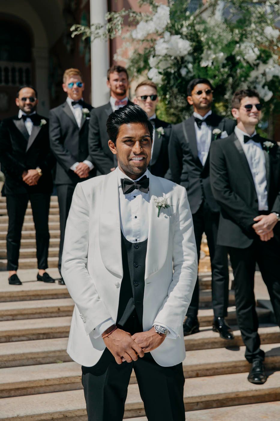 A group of groomsmen in formal black tuxedos and sunglasses stand on stone steps behind the groom in a white dinner jacket.