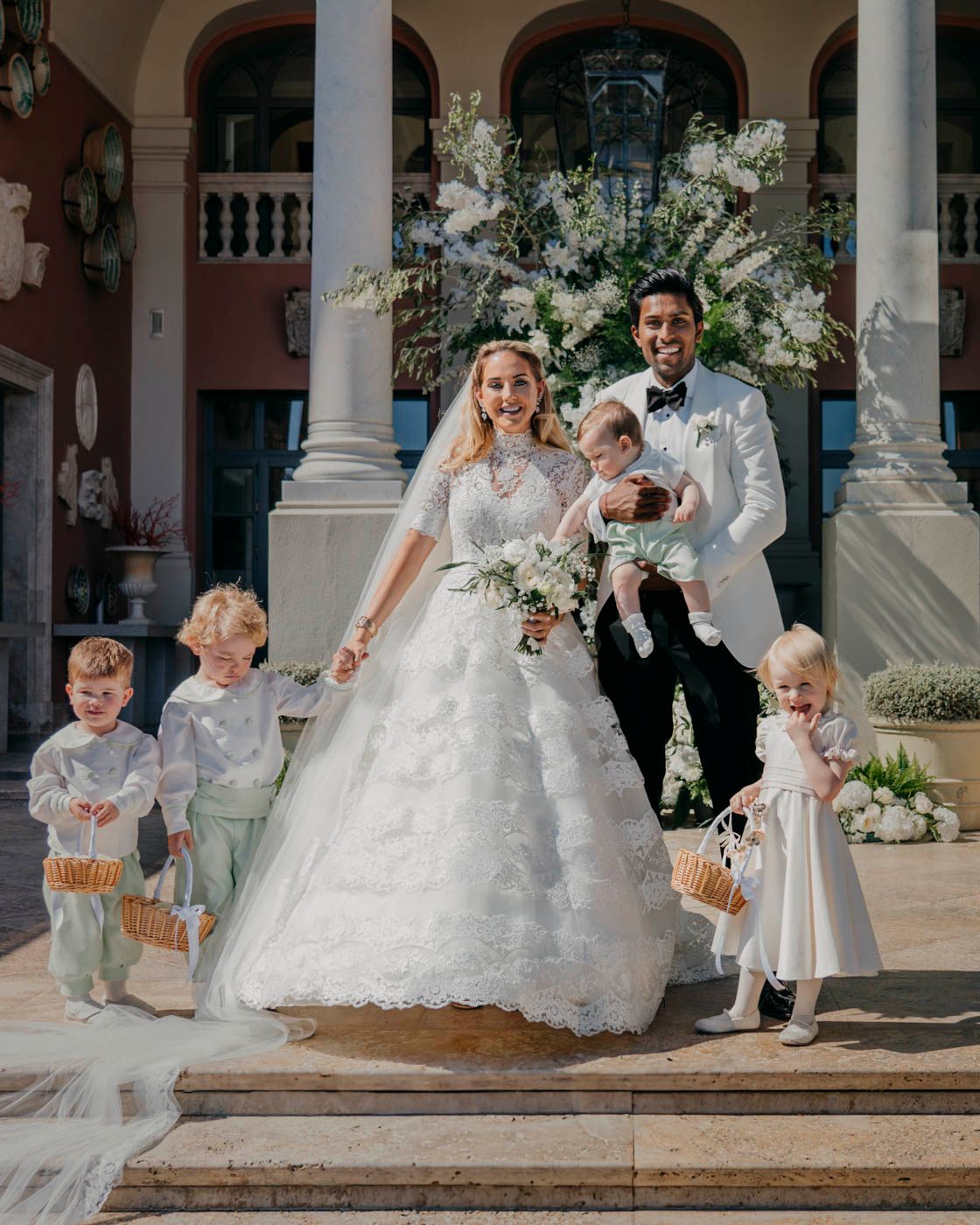 Wedding party stands on elegant mansion steps with lush floral arrangements and decorative white columns.