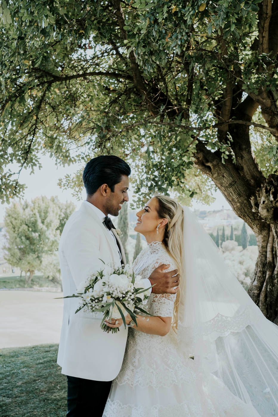 A romantic wedding portrait of a couple embracing under a tree, with the bride holding a white floral bouquet.