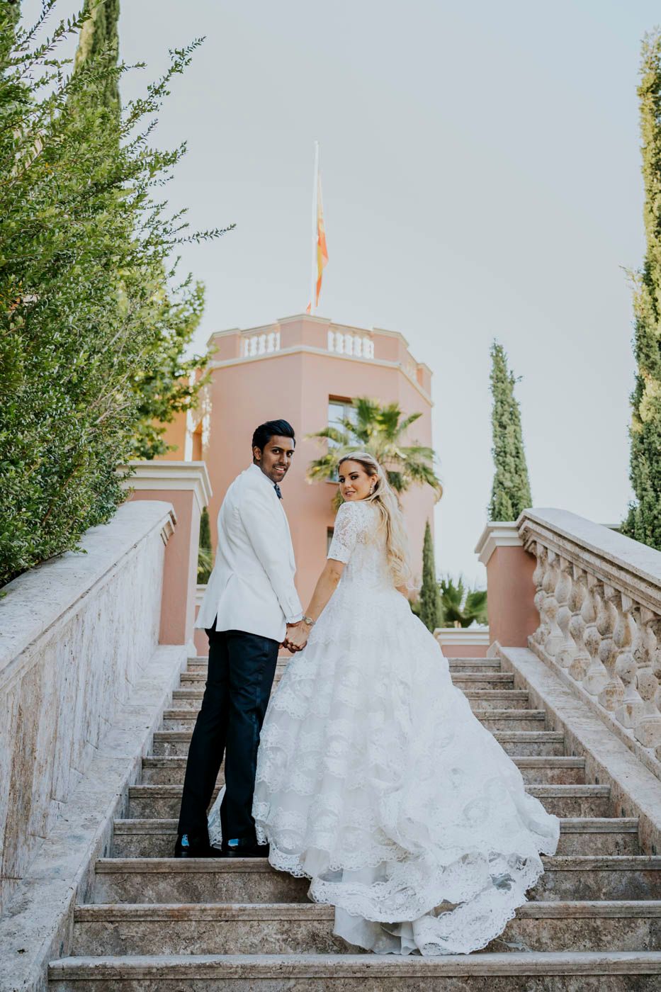 A newlywed couple poses on elegant stone steps with a pink Mediterranean-style building and cypress trees in background.