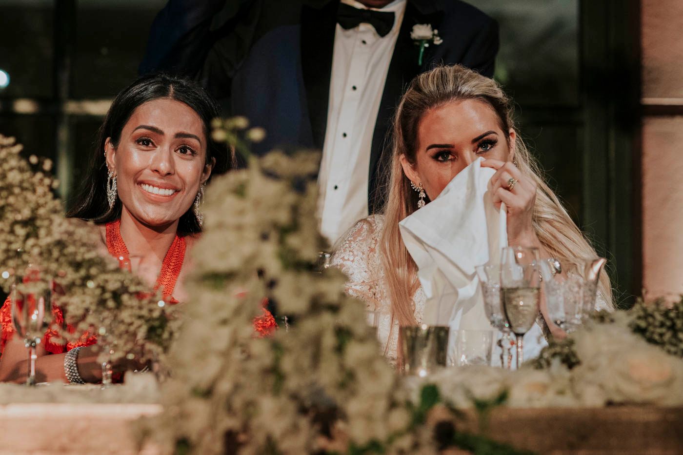 Two wedding guests share an emotional moment during a celebration while holding white napkins to their faces.
