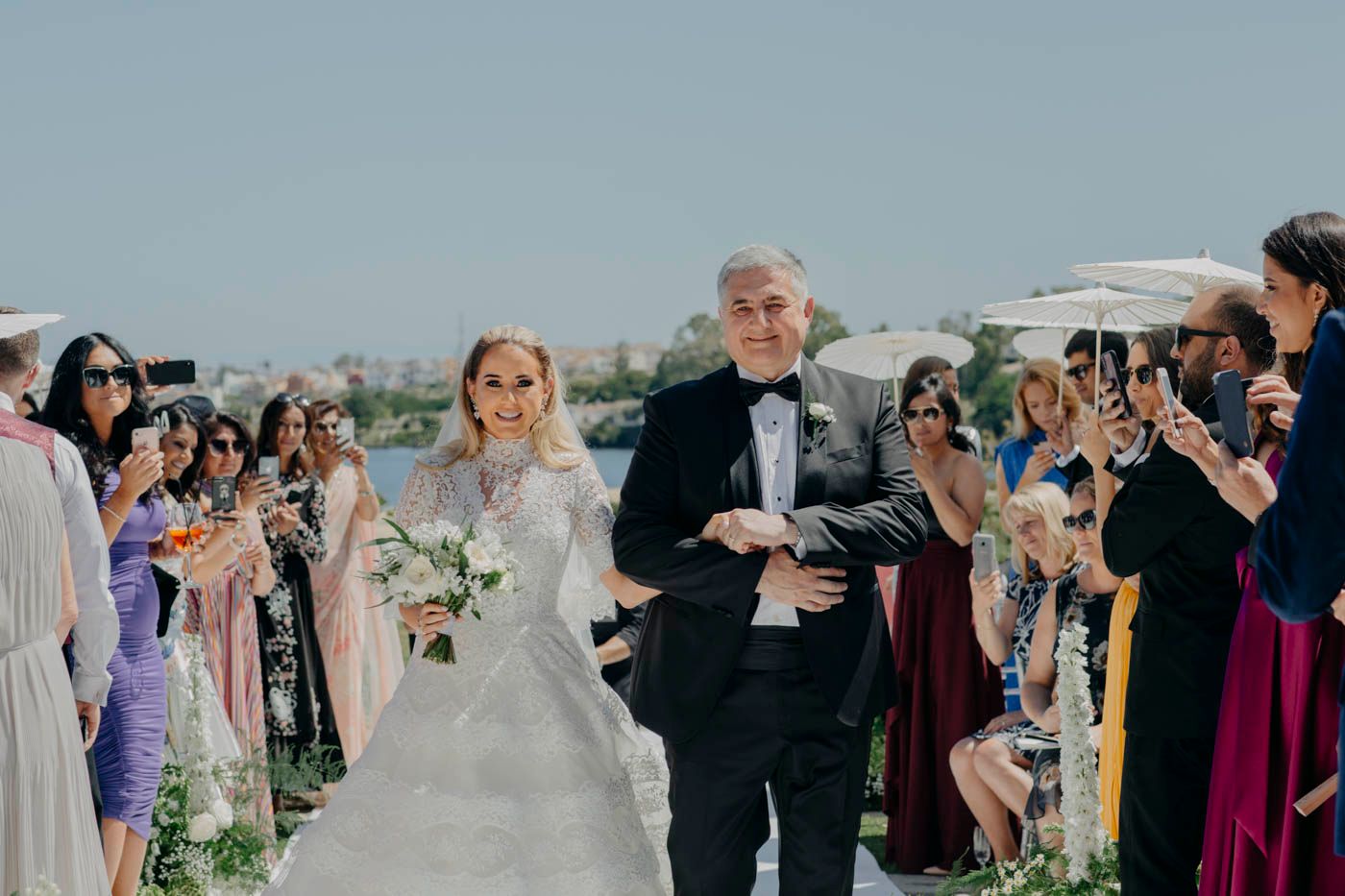 Wedding guests line an outdoor aisle while someone escorts someone in a white wedding gown during the ceremony.