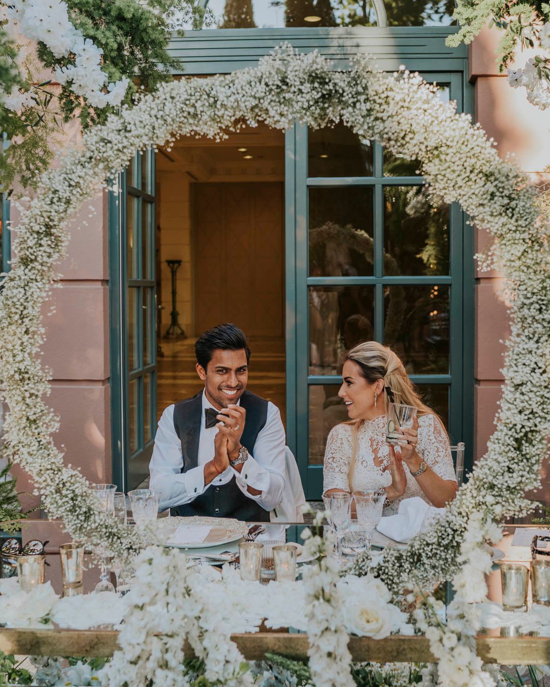 A couple shares a joyful moment at their wedding reception under an ornate white floral arch with glowing candles.
