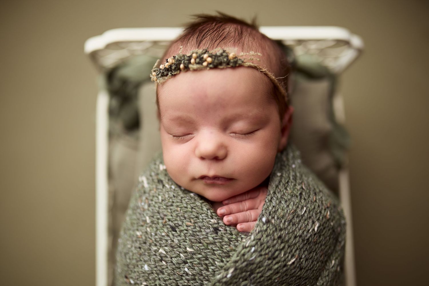 A sleeping newborn wrapped in a gray knit blanket wearing a delicate headband rests in a vintage chair.