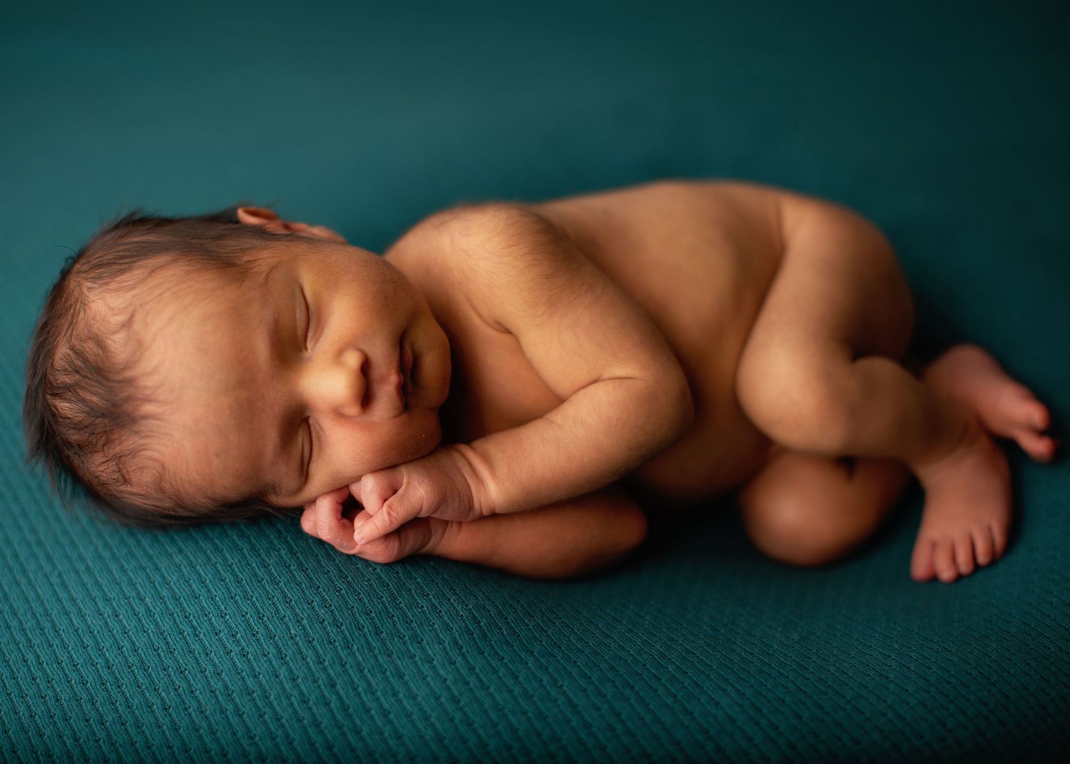 Peaceful sleeping newborn baby curled up on teal-colored textured backdrop in studio lighting.