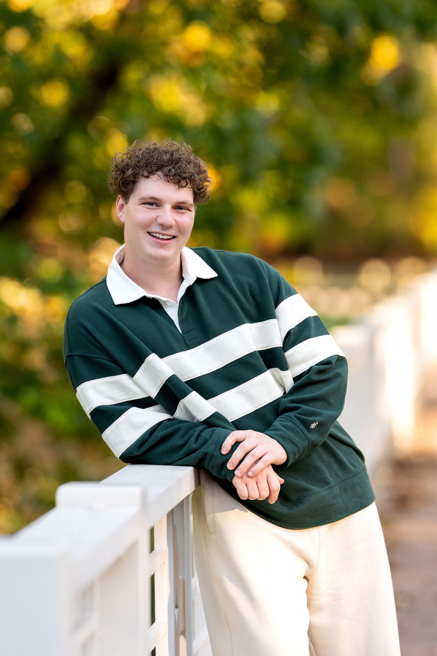 Smiling young man in green striped rugby shirt leaning on white fence with autumn foliage background.