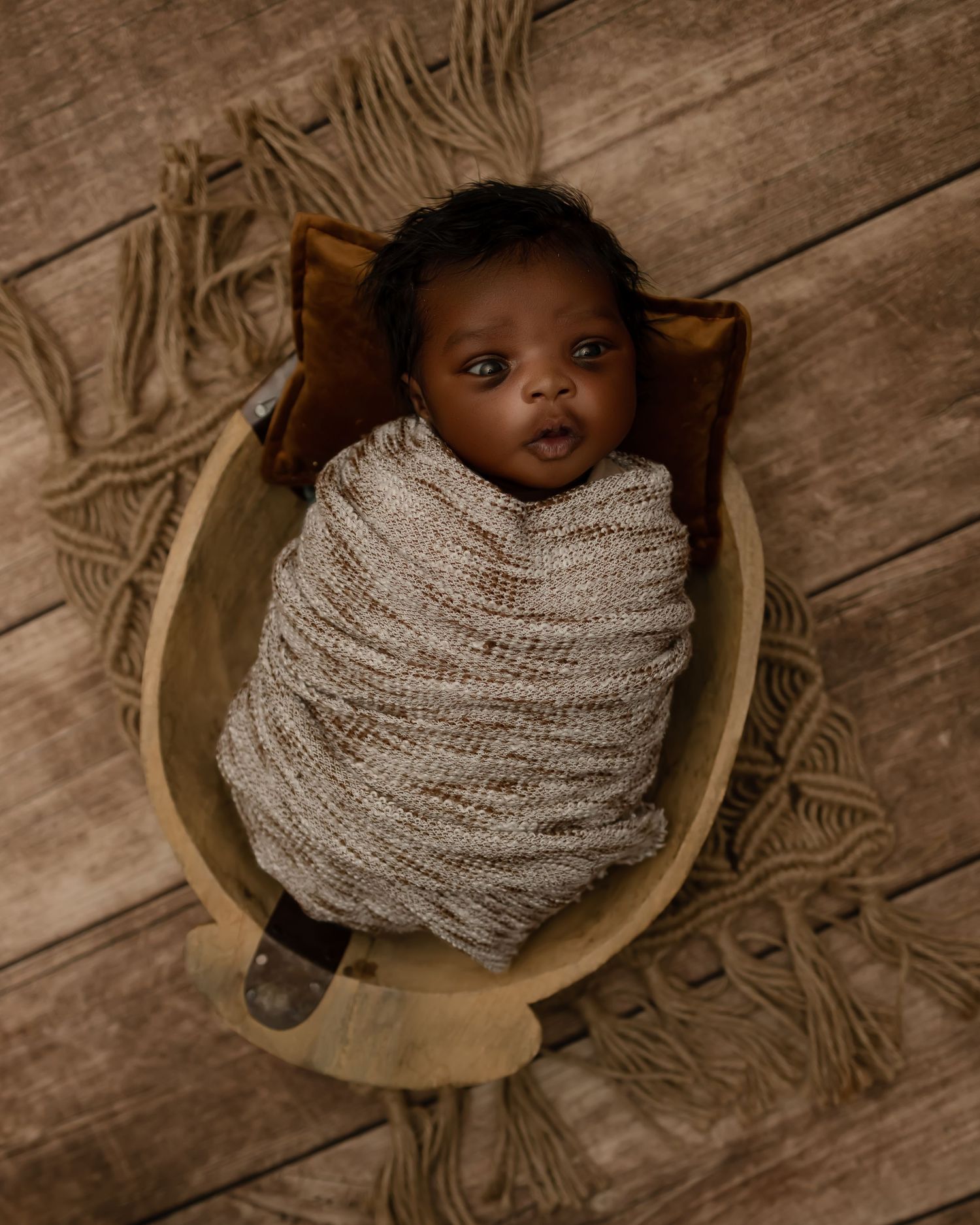 Small baby swaddled in cozy gray knit blanket lying in rustic wooden bowl against textured brown background.