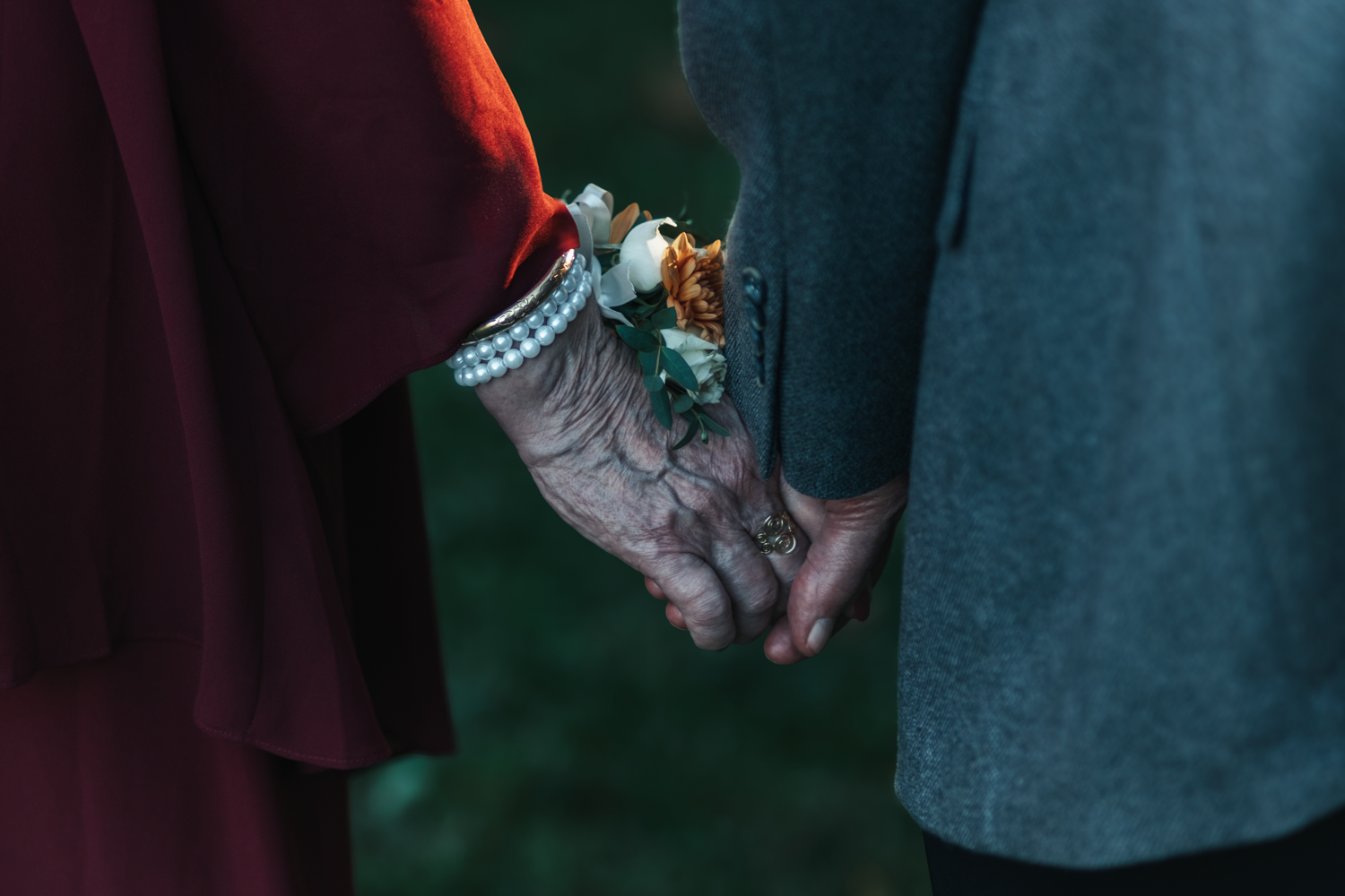 Close-up of two people holding hands with a delicate floral corsage between burgundy and teal colored fabrics.