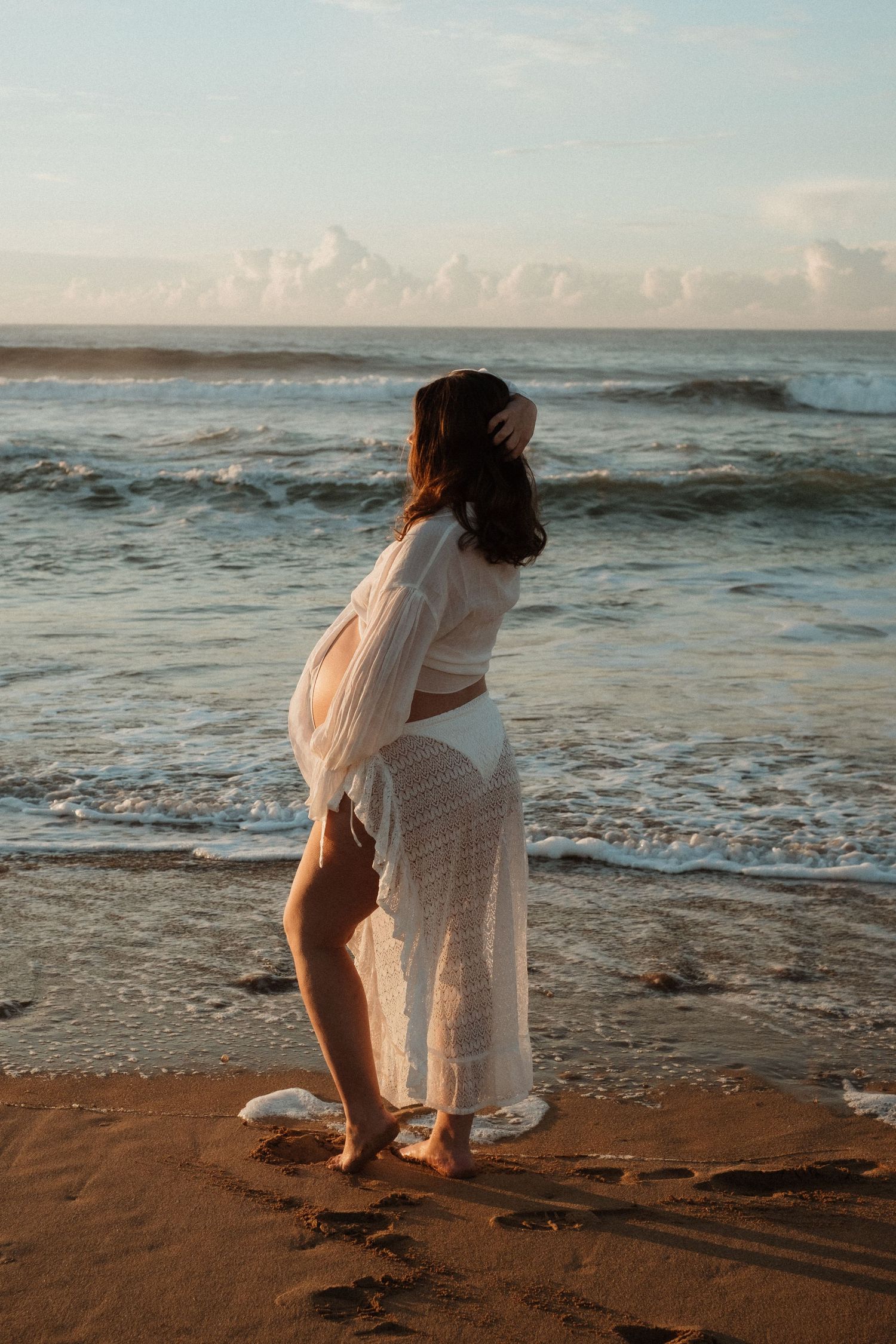 Expecting mother in flowing white dress gazing at ocean waves.