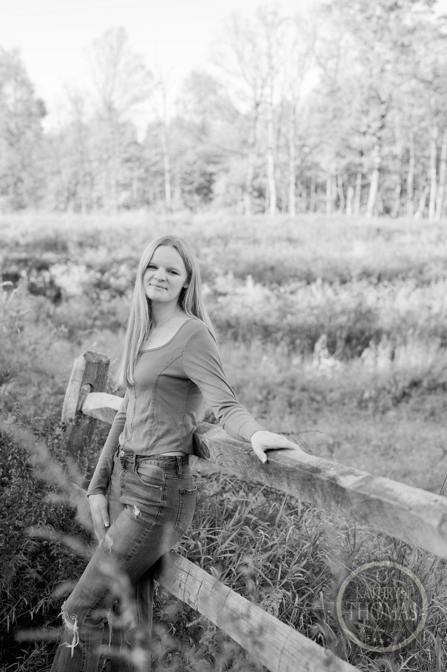 Black and white portrait showing someone leaning against wooden fence in natural outdoor setting.