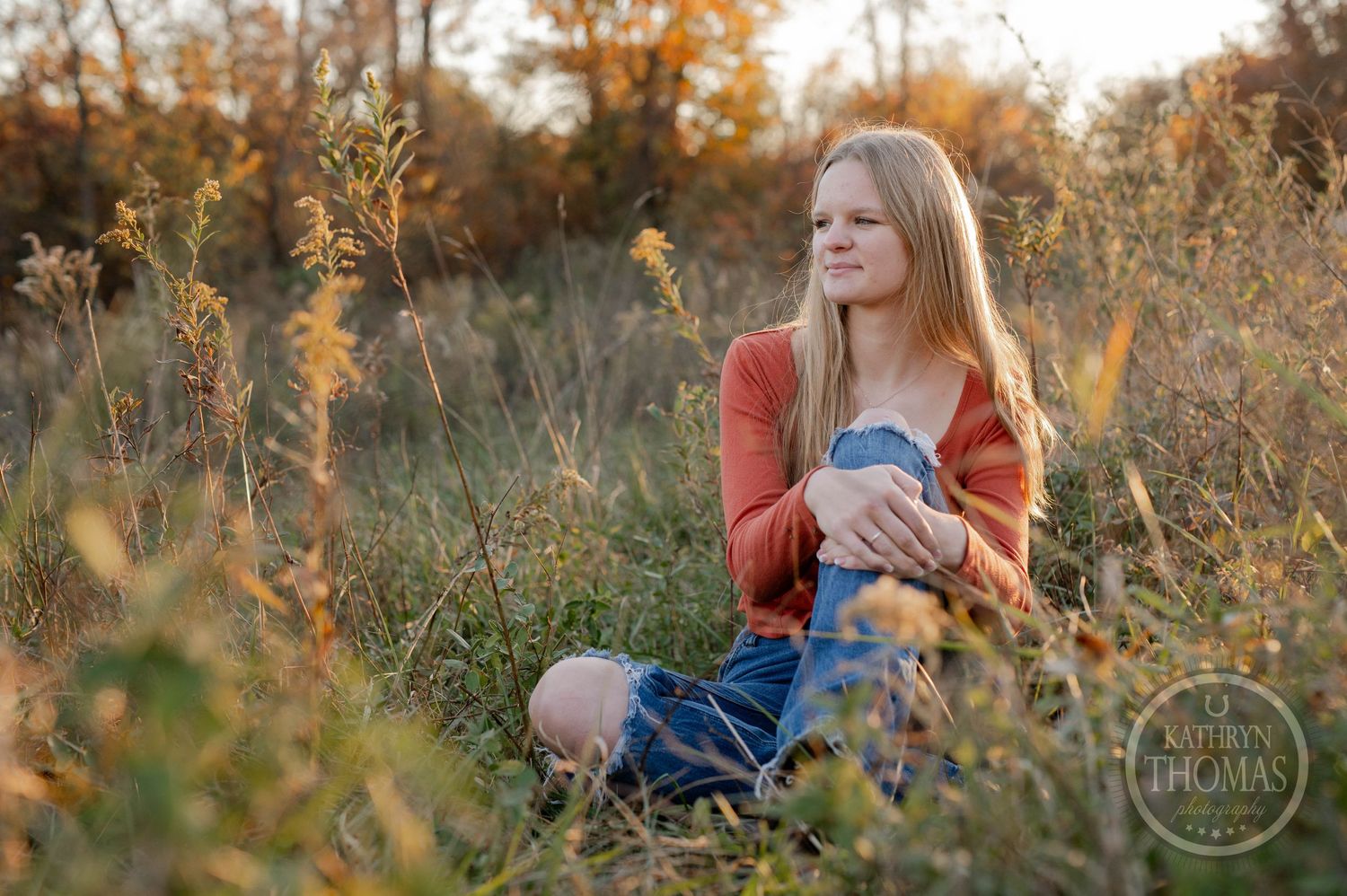 A person in an orange sweater sits in a golden autumn field at sunset among tall grass and wildflowers.