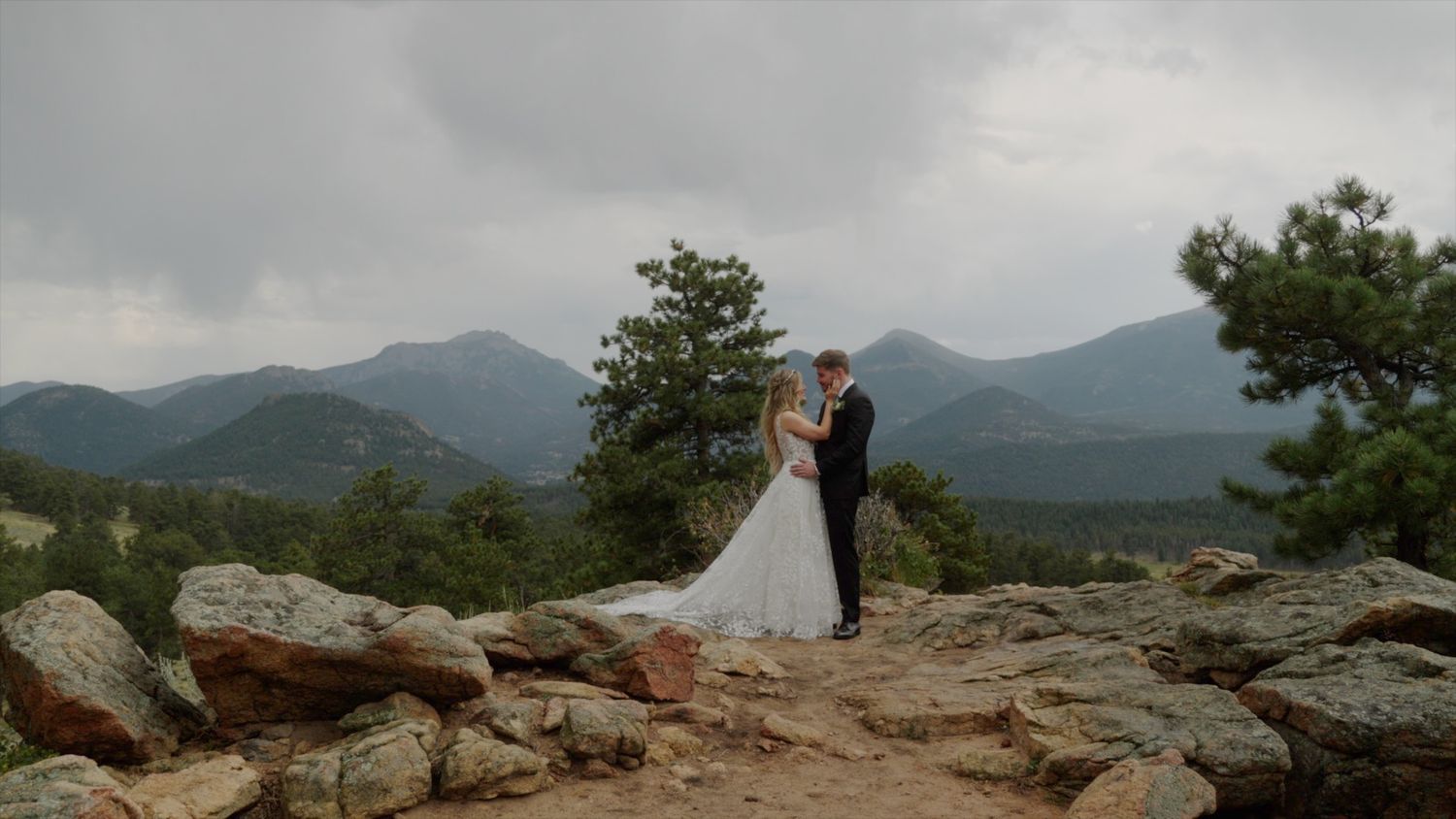 A couple shares a romantic moment on a rocky mountain overlook with misty peaks in the background.