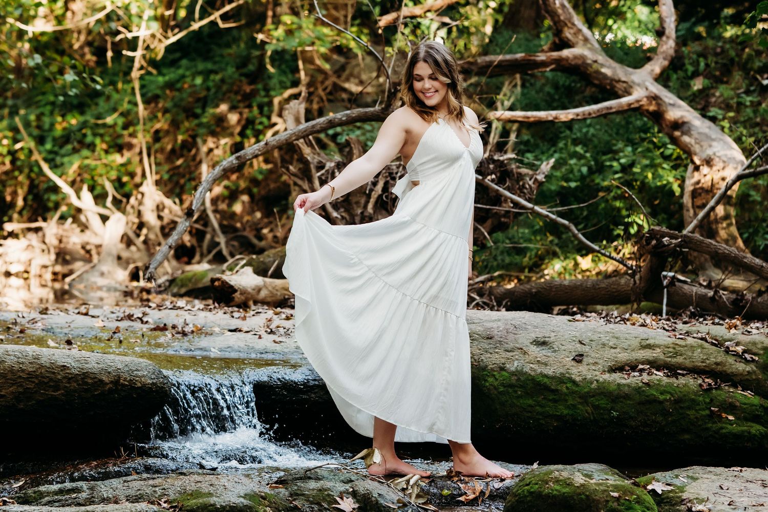 A person in a flowing white dress poses gracefully by a cascading waterfall in a lush forest setting.