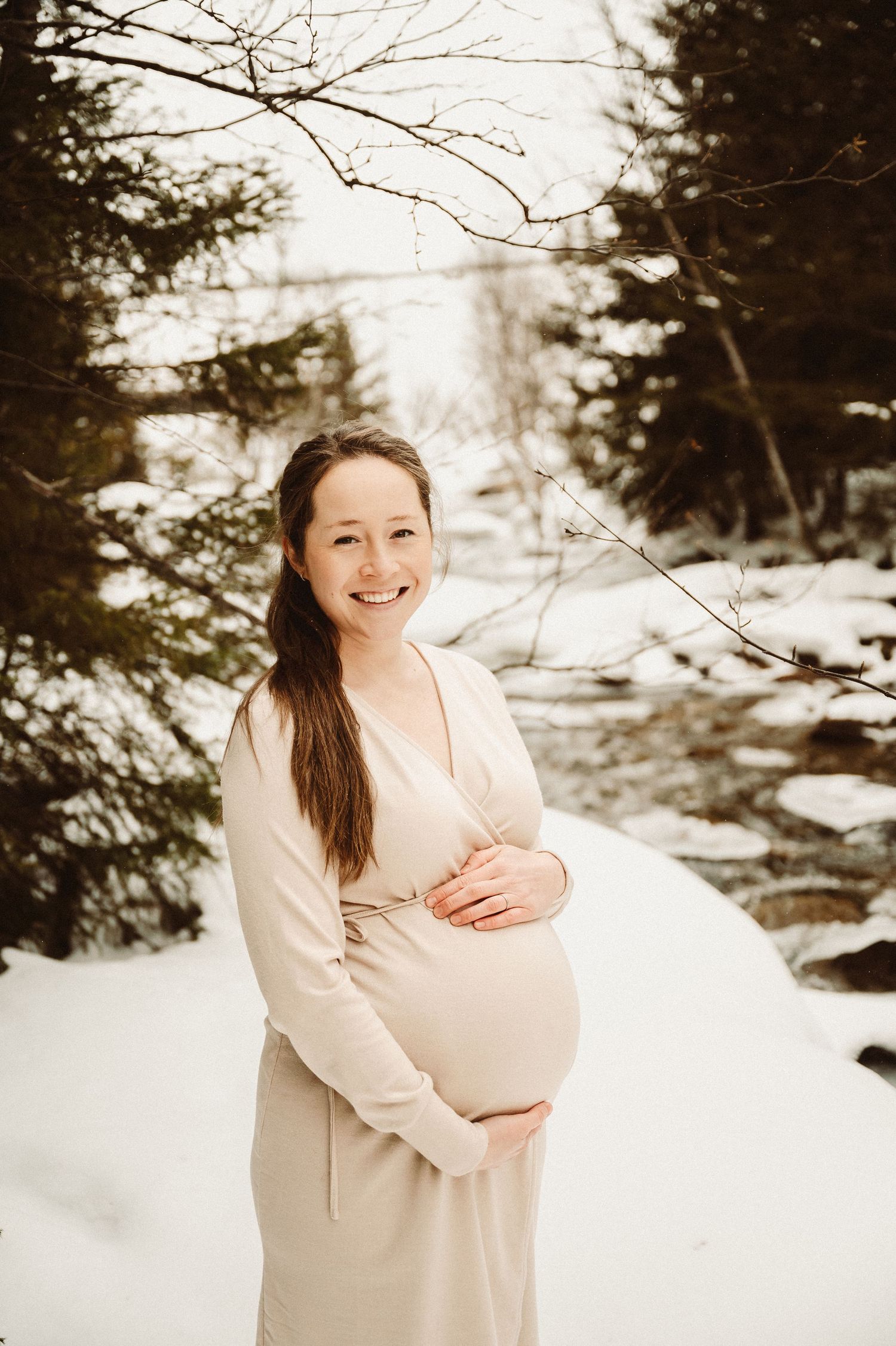 A maternity portrait in a snowy forest setting with winter trees framing the scene.