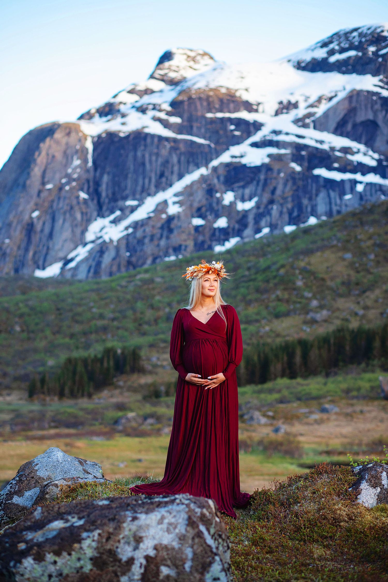 Pregnant woman in burgundy red gown with floral crown standing before snow-capped Norwegian mountains.
