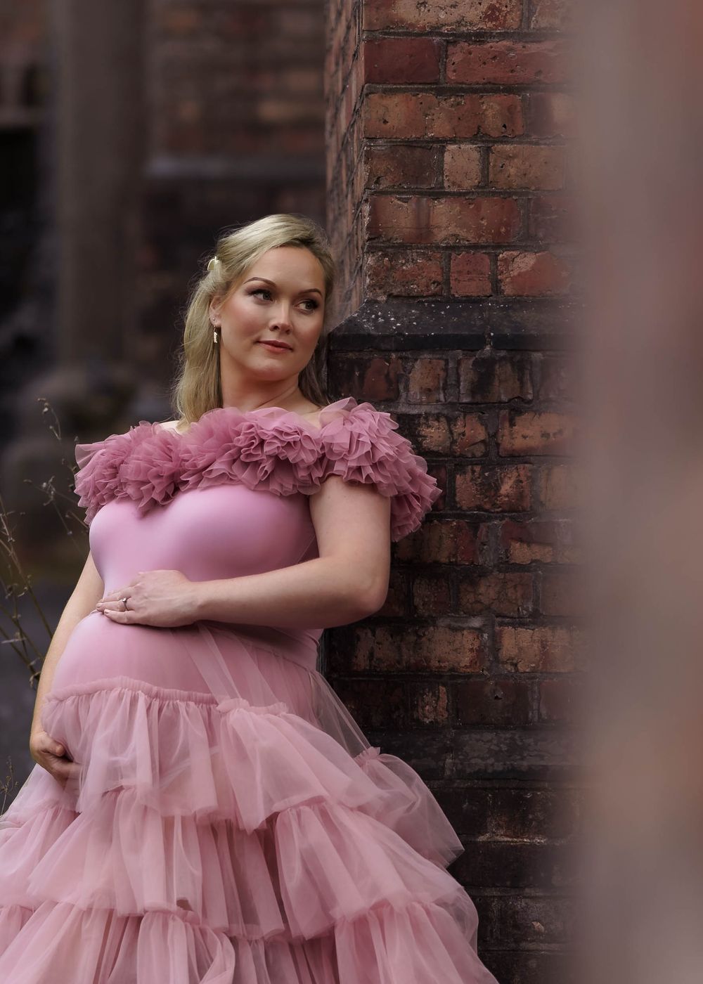 Pregnant woman in pink ruffled gown poses against brick wall in maternity photo.