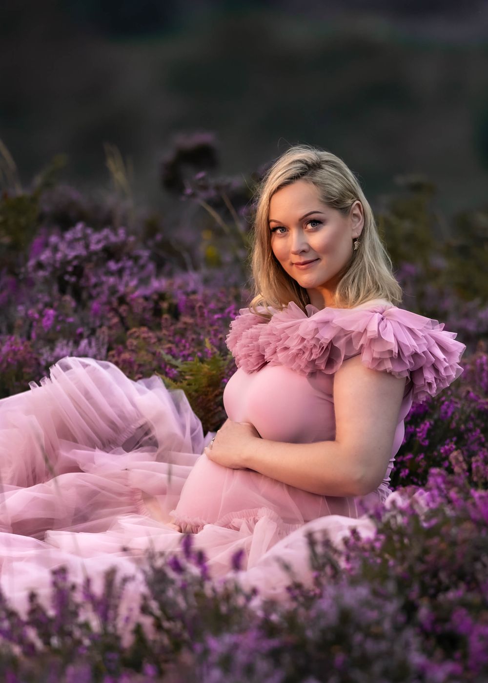 Blonde woman in pink ruffled gown poses for maternity photo among purple flowers.