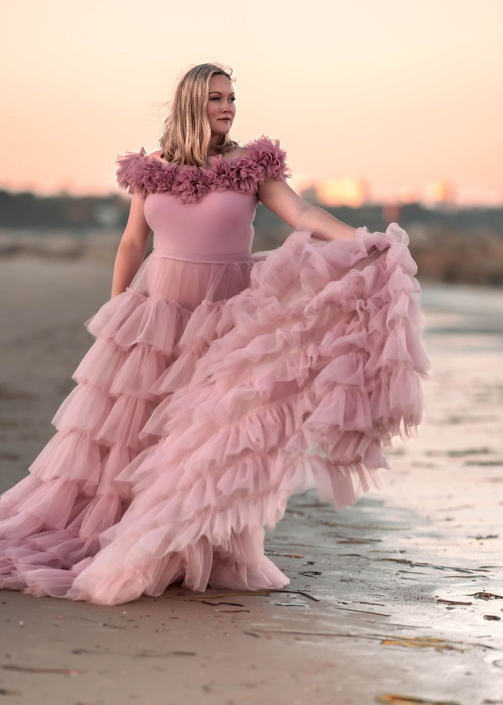 Pregnant woman in flowing pink gown stands on beach at sunset for maternity photo.