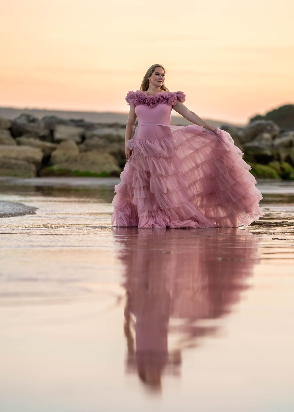 Dramatic maternity photo of woman in pink ruffled dress reflected in water at sunset.