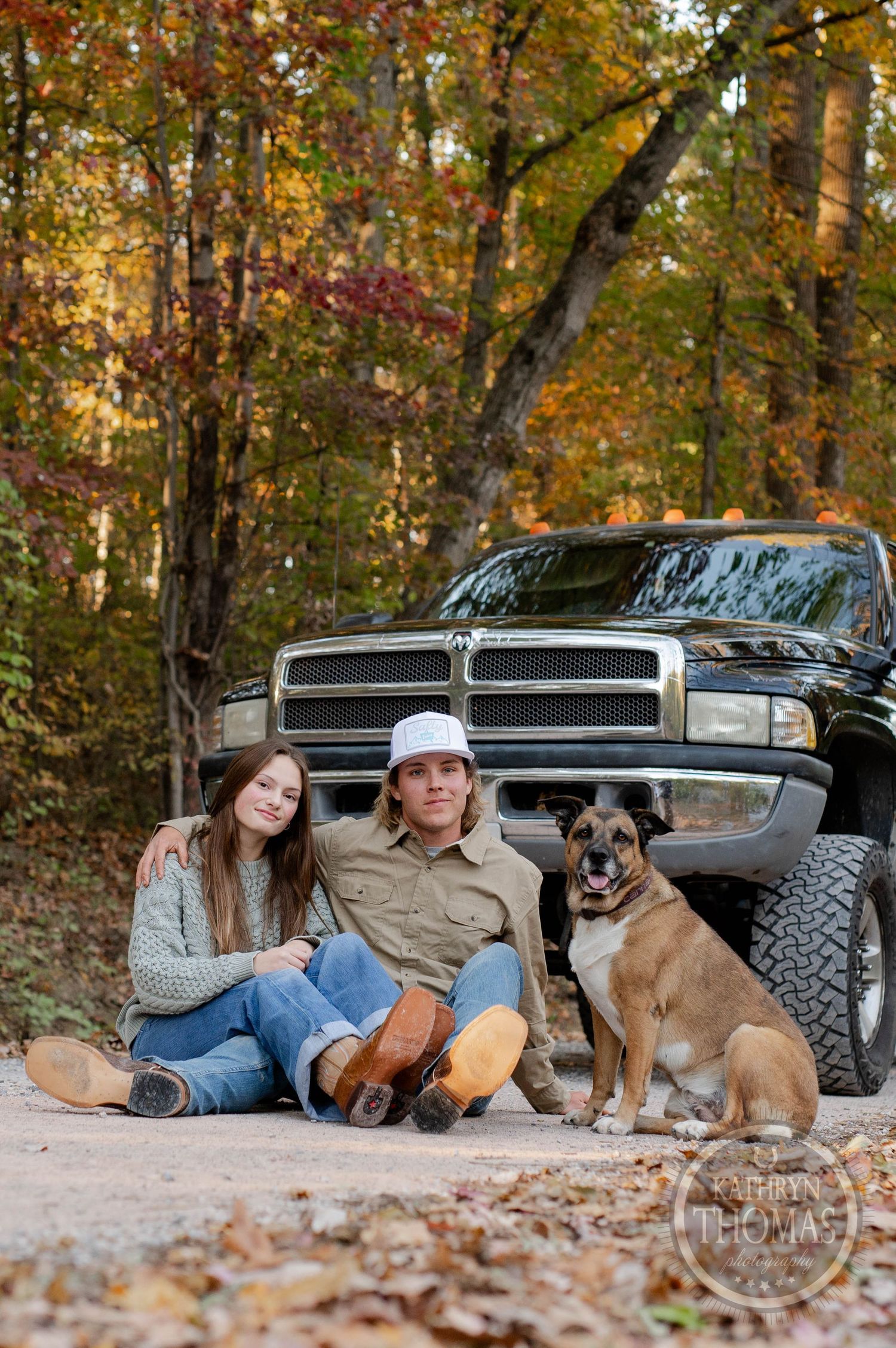 A brown dog sits with companions against a lifted black truck surrounded by fall foliage.