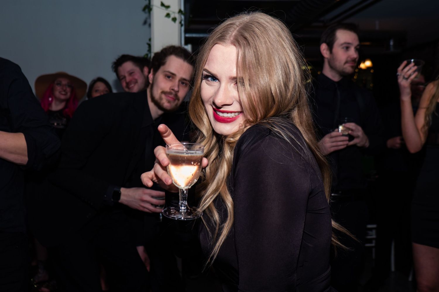 A person in a black outfit smiles while holding a cocktail glass at a dimly lit party event.