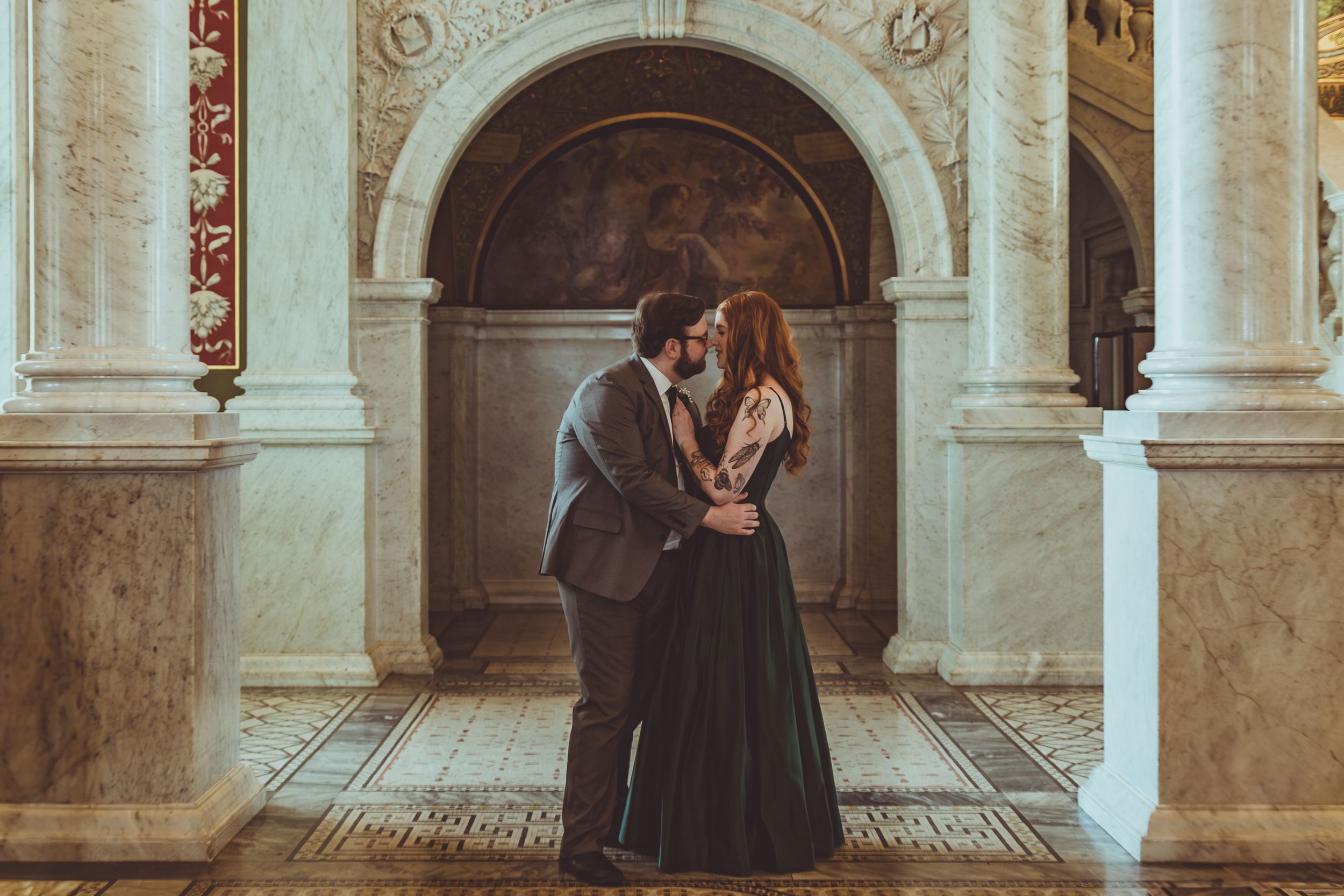 A romantic couple in formal attire embrace in front of an ornate archway in a vintage-inspired setting.