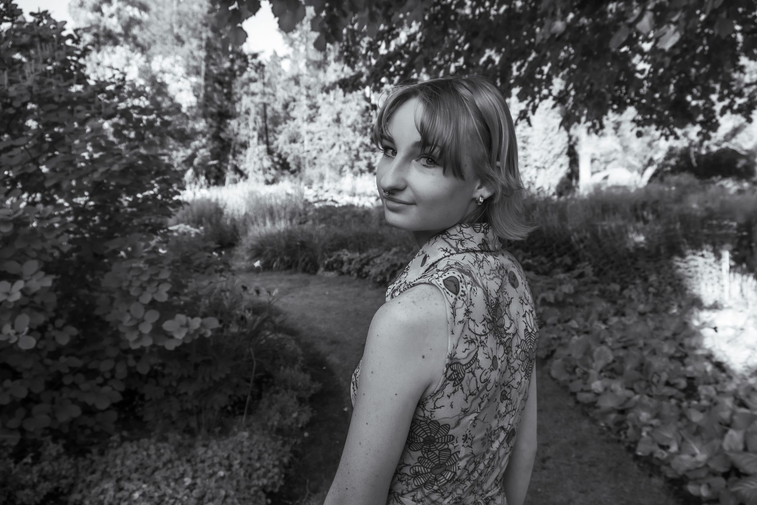 A black and white portrait photo shows a person in a sleeveless top looking back over their shoulder in a garden setting.