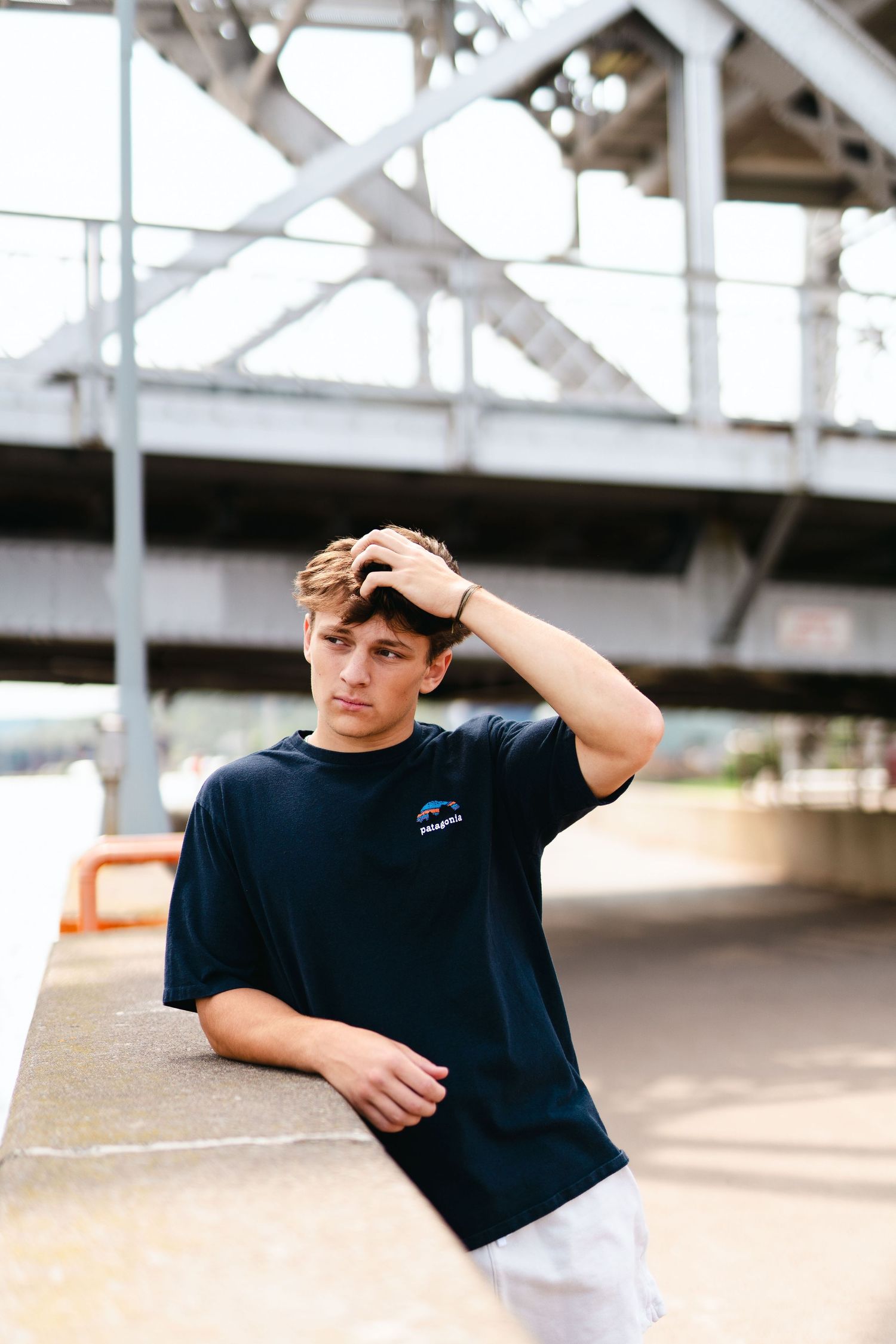 A person in a navy blue t-shirt poses under a bridge structure on a sunny day.